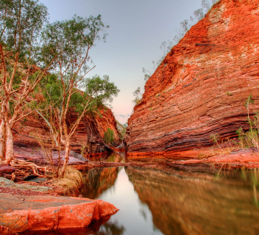 Two red and brown-streaked rocky gorges converge over a still pool of water, lined with trees and shrubs