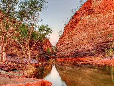 Two red and brown-streaked rocky gorges converge over a still pool of water, lined with trees and shrubs