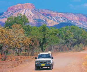 A four-wheel drive car drives down a red-dirt road, with the Pilbara mountain ranges in the distance
