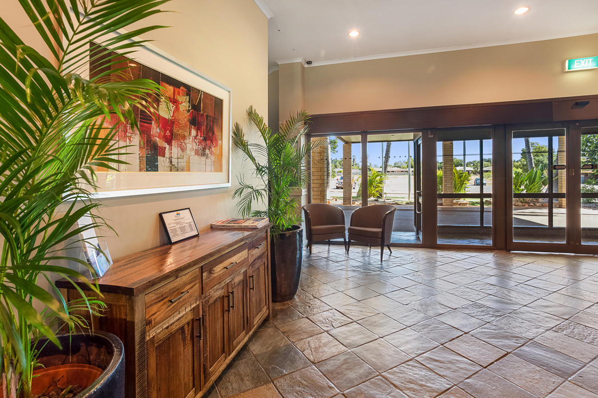 The Karratha International Hotel reception is decorated with stone tiling, wooden accent furniture, and potted palm plants.