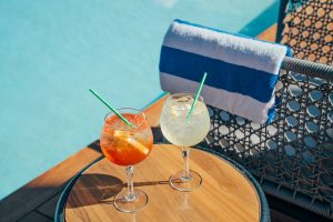 Two glasses of spritz cocktails are placed on a side table near a swimming pool