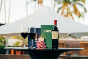 A cocktail table holds two glasses of red wine, a wine bottle, Luce Bar e Cibo menu and candle