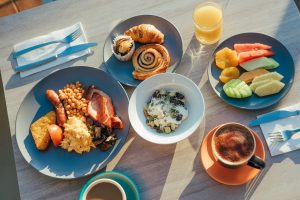 A restaurant table holds plates of breakfast dishes from Luce Bar e Cibo, including some pastries, fresh fruit, a cooked breakfast, granola and yogurt, orange juice and coffee