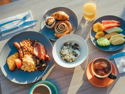 A restaurant table holds plates of breakfast dishes from Luce Bar e Cibo, including some pastries, fresh fruit, a cooked breakfast, granola and yogurt, orange juice and coffee