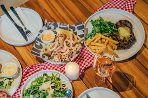 A restaurant table holds plates of Italian food, including some fried calamari rings, salad, steak and fries